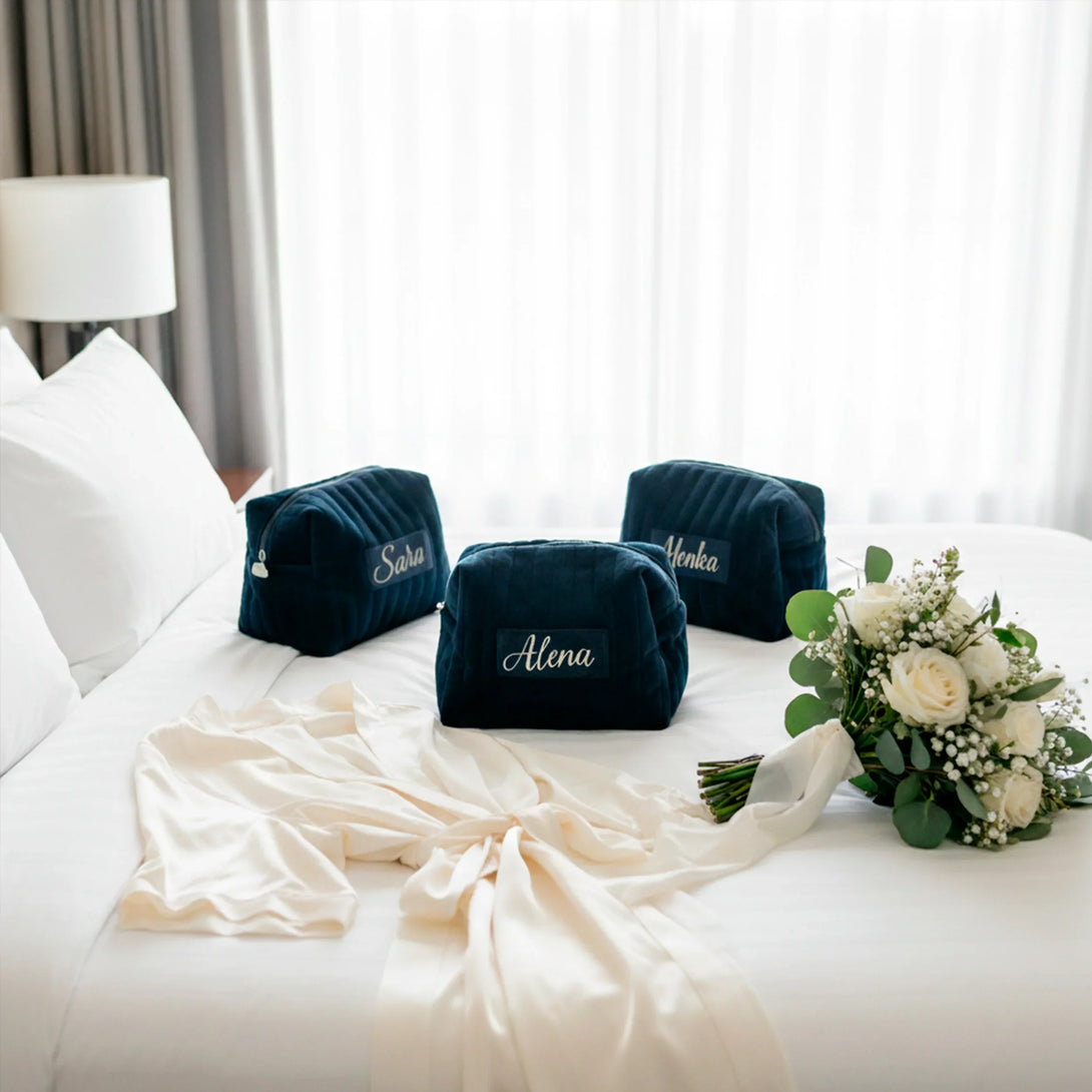 Three personalized navy blue velvet pouches with female names on a white bed beside a cream satin robe and white flower bouquet